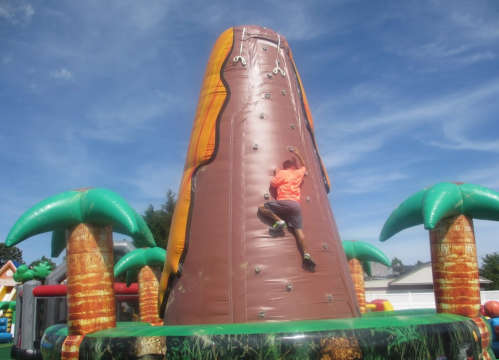 A person climbs a large inflatable volcano surrounded by palm trees under a blue sky.