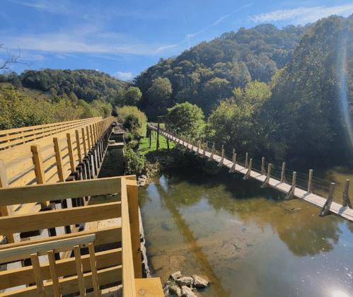 A scenic view of two wooden bridges crossing over a calm river, surrounded by lush green hills under a clear blue sky.