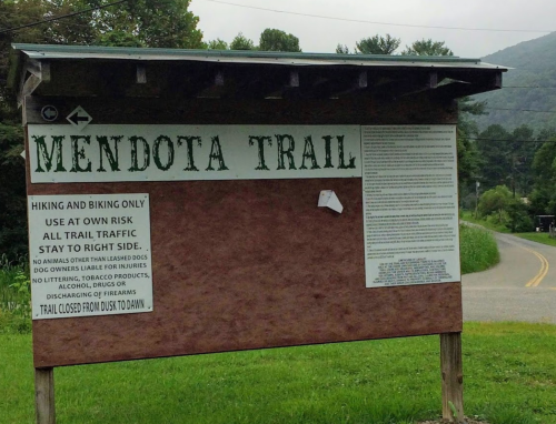 Sign for Mendota Trail with hiking and biking rules, located near a road surrounded by trees and mountains.