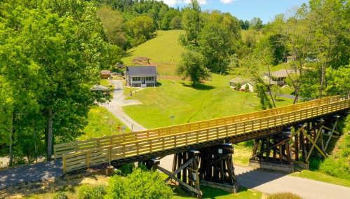 A wooden bridge spans over a road, surrounded by lush green fields and trees in a rural landscape.