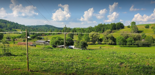 A lush green landscape with rolling hills, trees, and a blue sky dotted with fluffy clouds. Fenced pasture in the foreground.