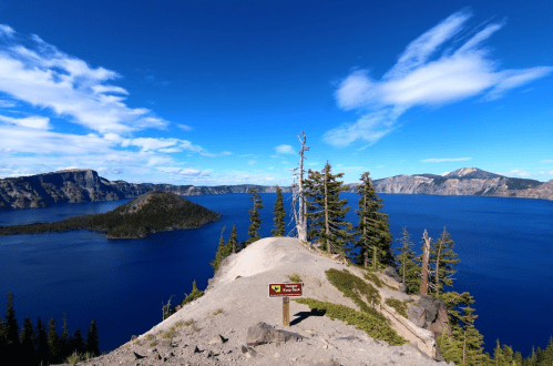 A scenic view of Crater Lake with blue water, trees, and a signpost against a clear sky.