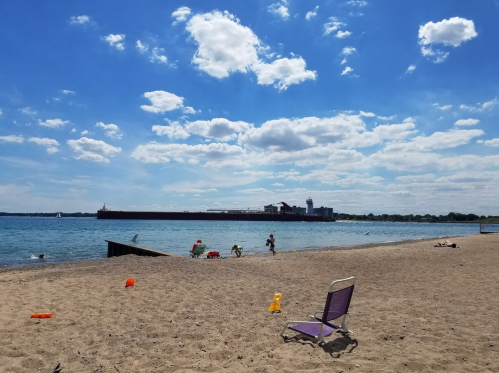 A sunny beach scene with a sandy shore, people enjoying the water, and a distant pier under a blue sky with clouds.
