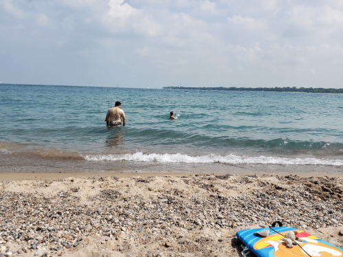 A man wades in shallow water while a child swims nearby, with a colorful beach mat on the sandy shore.
