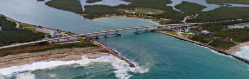 Aerial view of a bridge over turquoise waters, connecting land with lush greenery and sandy beaches.