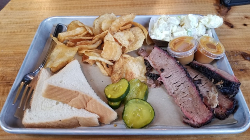 A metal tray with sliced brisket, pickles, potato chips, bread, and two containers of sauce.
