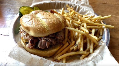 A pulled pork sandwich on a bun with fries and pickles on the side, served in a metal tray.
