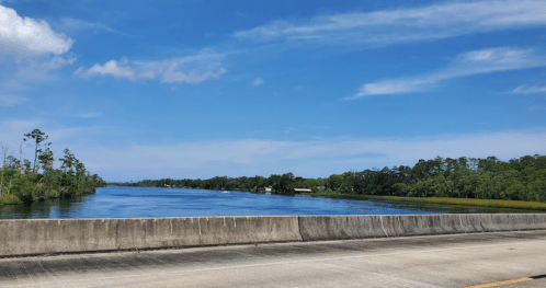A serene river scene with blue skies, green trees, and a calm waterway, viewed from a roadside.