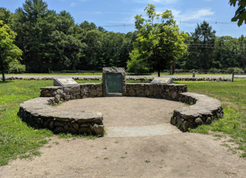 A circular stone monument in a grassy area, surrounded by trees, with informational plaques on either side.