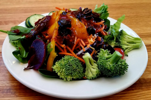 A colorful salad with mixed greens, carrots, black beans, broccoli, cucumbers, and a tangy dressing on a white plate.