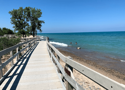 A wooden boardwalk leads to a beach with two people in the water, surrounded by trees and a clear blue sky.