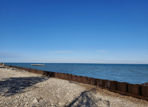 A calm shoreline with a wooden barrier, pebbles, and a clear blue sky over the water.