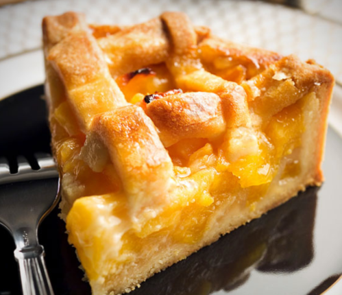 A slice of golden peach pie with a lattice crust, served on a black plate with a fork beside it.