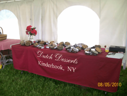 A table with a red tablecloth displaying various Dutch desserts at an outdoor event under a tent.