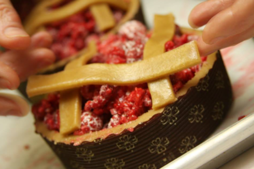 A close-up of hands placing a lattice crust on a raspberry pie filling in a decorative pie dish.