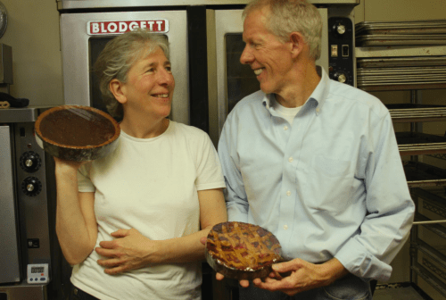 Two smiling people hold pies in a kitchen, standing in front of ovens and baking trays.