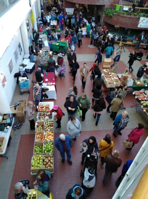 A bustling indoor market with vendors and shoppers, featuring tables of produce and a crowd of people browsing.