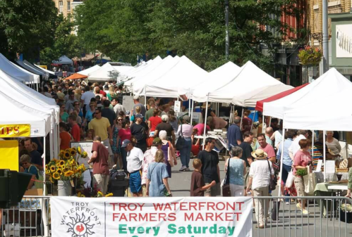 A bustling farmers market with numerous tents, people browsing, and vibrant greenery in the background.