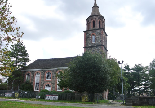 Historic church with a tall steeple, surrounded by trees and a grassy area, under a cloudy sky.