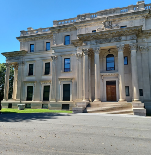 A grand, historic mansion with large columns and a staircase, set against a clear blue sky.