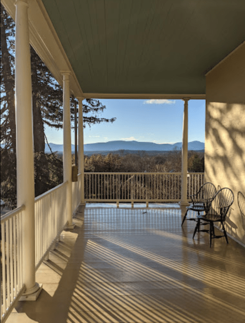 A porch with white railings and chairs, overlooking a scenic view of mountains and trees under a clear blue sky.