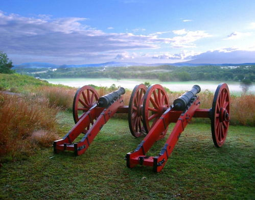 Two red cannons on a grassy field, overlooking a misty landscape with mountains and a cloudy sky in the background.