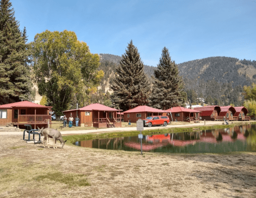 A scenic view of wooden cabins by a pond, surrounded by trees and mountains, with people and a deer nearby.