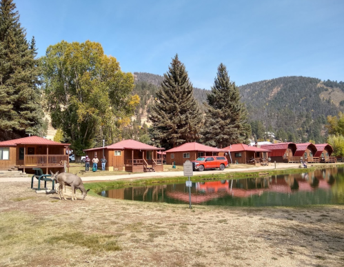 A scenic view of wooden cabins by a pond, surrounded by trees and mountains, with people and a deer nearby.