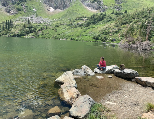 A person sits on rocks by a serene lake, surrounded by lush green hills and trees under a clear sky.