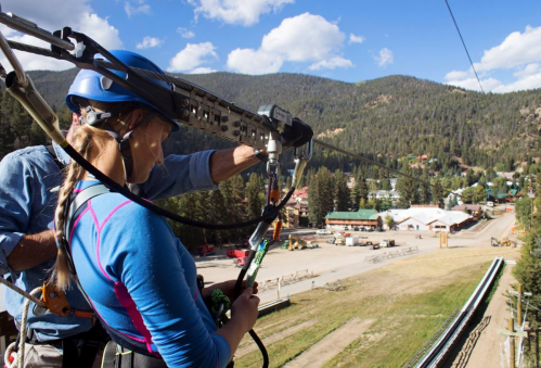 A person prepares to zip line, wearing a helmet and harness, with a scenic mountain view and a small town below.