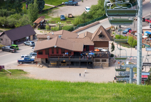 Aerial view of a rustic building with outdoor seating, surrounded by greenery and a ski lift in the foreground.