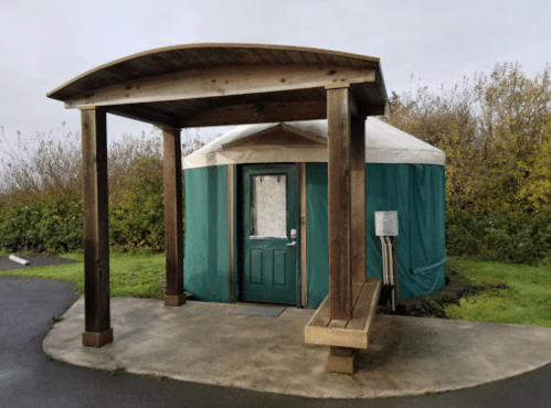 A green yurt with a wooden entrance and bench, surrounded by greenery and a cloudy sky.