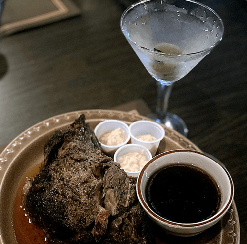 A plate of roasted meat with dipping sauce and sides, accompanied by a martini in a glass.