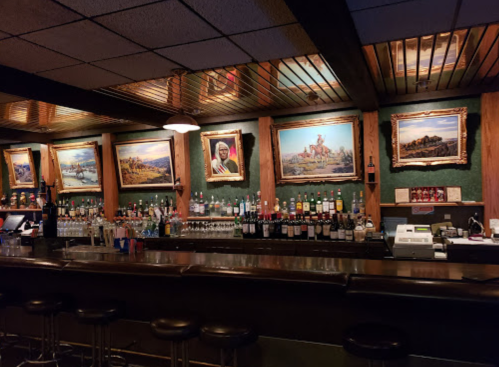 A cozy bar with a wooden counter, shelves of liquor, and framed landscape paintings on the walls.