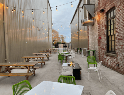 Outdoor seating area with string lights, picnic tables, and a fire pit, surrounded by industrial-style buildings.