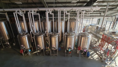 A row of large stainless steel brewing tanks in a modern brewery, with pipes and equipment in the background.