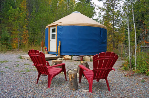 A blue yurt surrounded by trees, with two red chairs and a small fire pit in front on a gravel area.