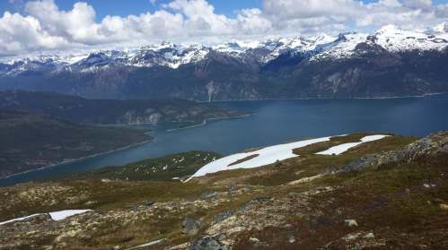 A panoramic view of snow-capped mountains and a serene blue lake under a partly cloudy sky.
