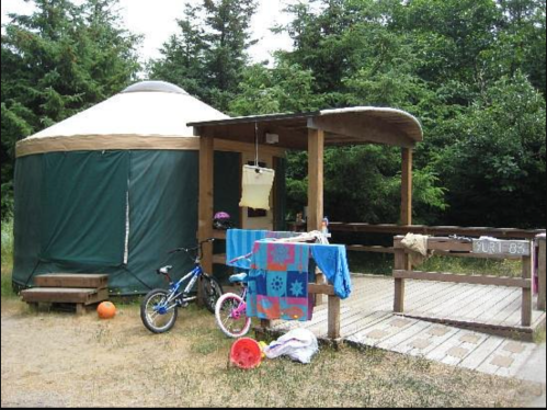 A yurt with a wooden deck, surrounded by trees, featuring a bike, toys, and towels hanging on a railing.
