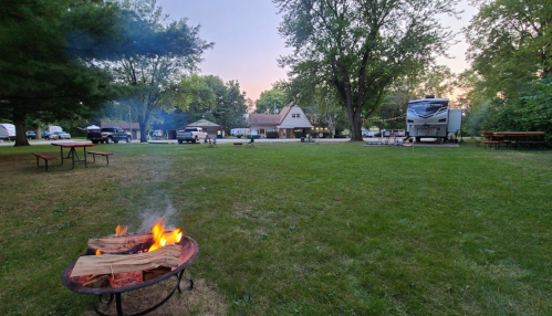 A campfire burns in the foreground, with RVs and picnic tables in a grassy area under a twilight sky.
