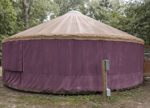 A round yurt with a tan roof and purple fabric walls, set in a wooded area.
