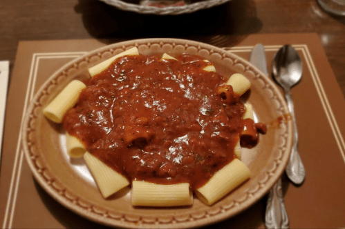 A plate of rigatoni pasta topped with a rich meat sauce, served on a decorative dish with silverware beside it.