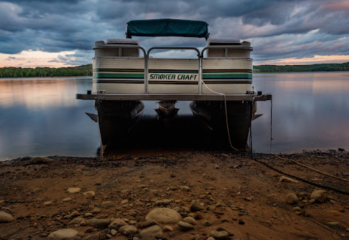 A pontoon boat on a calm lake shore under a cloudy sky, with trees in the background and a rocky foreground.