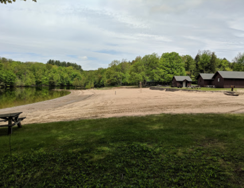 A sandy beach by a calm lake, surrounded by green trees and rustic cabins in the background.