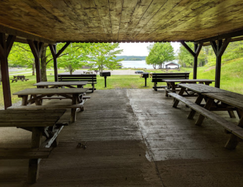 View from a covered picnic area with wooden tables, overlooking a grassy area and a lake in the background.