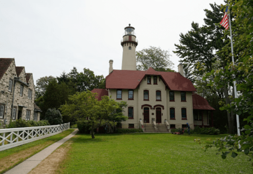 A historic lighthouse next to a large, two-story house, surrounded by greenery and a flagpole with an American flag.