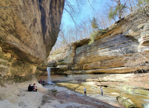 A scenic canyon with a waterfall, surrounded by rocky cliffs and trees, with people exploring the area.