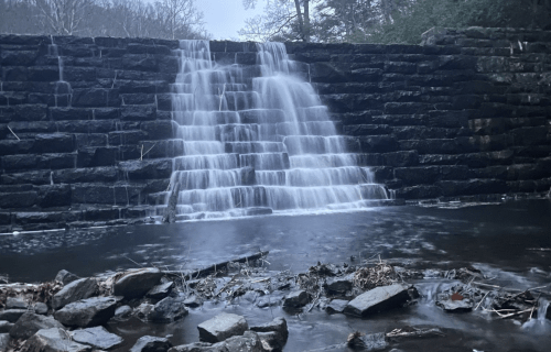 A serene waterfall cascading over a stone wall into a calm pool, surrounded by rocks and trees in a misty atmosphere.