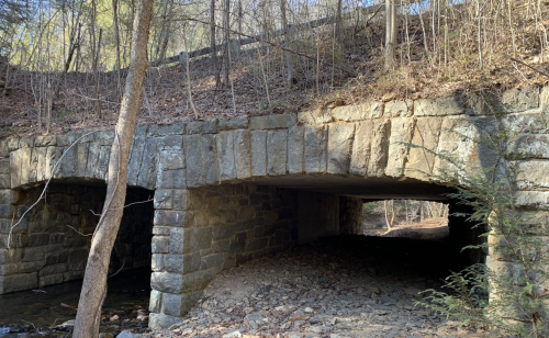 Stone arch bridge with two openings, surrounded by trees and rocky terrain, under a clear blue sky.