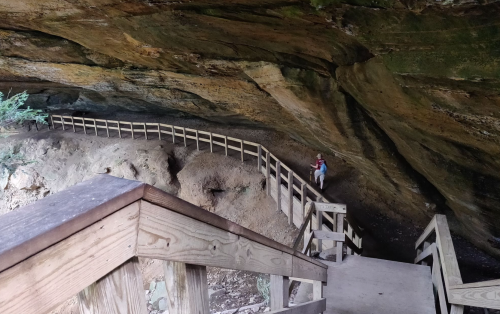 A wooden staircase leads down into a cave with a winding path and a person standing in the distance.